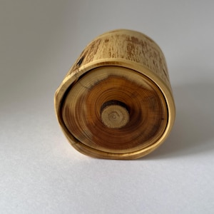 May include: A cylindrical wooden container with a natural bark exterior and a circular lid. The lid features a prominent central wooden knob, showcasing visible concentric growth rings in shades of light and dark brown wood grain.