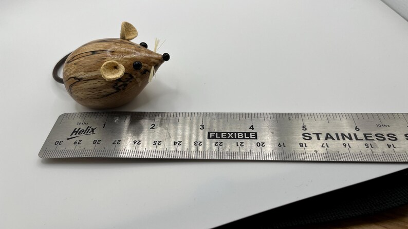 Puede incluir: Una peque&ntilde;a figura de rat&oacute;n de madera redondeada con acabado de veta natural, dos orejas peque&ntilde;as, ojos de cuentas negras y bigotes finos. Una regla de acero inoxidable Helix debajo muestra una longitud de aproximadamente 3,8 cm.