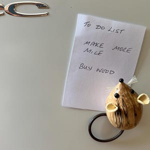 Puede incluir: Una nota rectangular blanca con el texto escrito a mano TO DO LIST, MAKE MORE MILE y BUY WOOD, sujeta por un im&aacute;n con forma de rat&oacute;n marr&oacute;n texturizado con ojos negros, nariz negra, bigotes blancos y una cola negra curvada sobre una superficie.