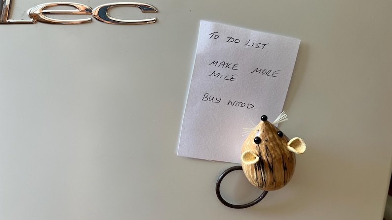 Puede incluir: Una nota rectangular blanca con el texto escrito a mano TO DO LIST, MAKE MORE MILE y BUY WOOD, sujeta por un im&aacute;n con forma de rat&oacute;n marr&oacute;n texturizado con ojos negros, nariz negra, bigotes blancos y una cola negra curvada sobre una superficie.