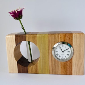 May include: A handcrafted wooden vase and clock combination. The vase features a circular opening for a flower and a quartz clock with Roman numerals. The wood is a mix of light and dark tones, with a purple and white flower.