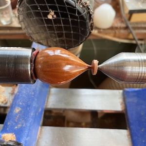 Puede incluir: Un objeto de madera pulida en forma de l&aacute;grima con un acabado suave y brillante y veta visible, asegurado horizontalmente entre dos centros de torno de metal. La pieza presenta un cuerpo bulboso redondeado que se estrecha en un cuello fino.