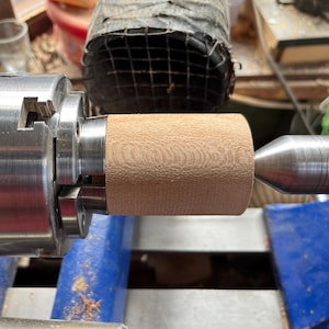Puede incluir: Una pieza cil&iacute;ndrica de madera clara est&aacute; asegurada en un mandril de torno de metal a la izquierda y apoyada por un contrapunto de metal a la derecha. La superficie de la madera muestra un grano fino y texturizado del proceso de torneado.