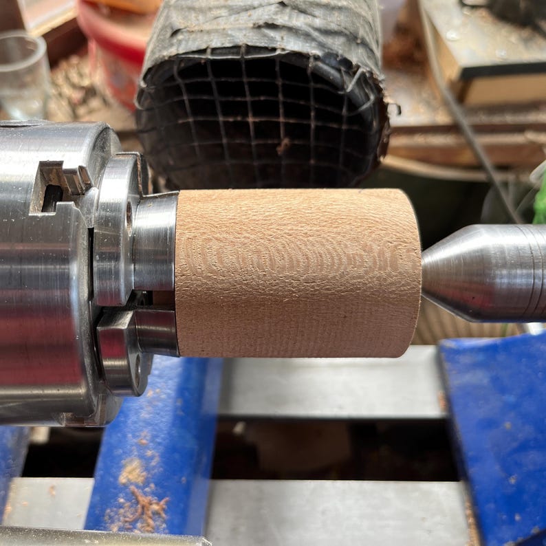 Puede incluir: Una pieza cil&iacute;ndrica de madera clara est&aacute; asegurada en un mandril de torno de metal a la izquierda y apoyada por un contrapunto de metal a la derecha. La superficie de la madera muestra un grano fino y texturizado del proceso de torneado.