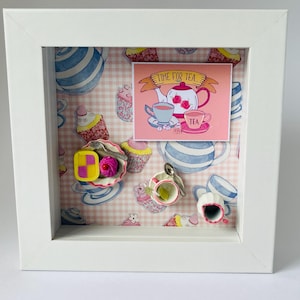 Op de afbeelding: Een wit omkaderde schaduwdoos met een theefeestthema. De achtergrond heeft een roze en wit geblokt patroon met illustraties van theekopjes en cupcakes. Een klein kaartje leest "Time for Tea" met een theepot en kopjes.