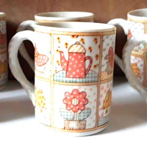 May include: Set of six ceramic mugs with white handles. Each mug features a patchwork design with a red teapot, a flower, and other decorative elements. The mugs have a cream-colored background with a floral pattern.