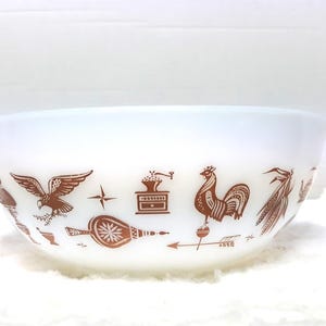 May include: A white milk glass mixing bowl with two side handles, featuring a repeating brown graphic pattern of an eagle, a coffee grinder, a rooster, a bellows, and wheat stalks printed around the exterior circumference of the bowl.