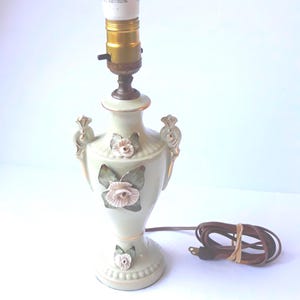May include: A pale green ceramic table lamp with a gold-trimmed urn shape, featuring three-dimensional pink rose appliqués with green leaves. The lamp has a brass-colored socket, a white light bulb, and a brown electrical cord with a plug.