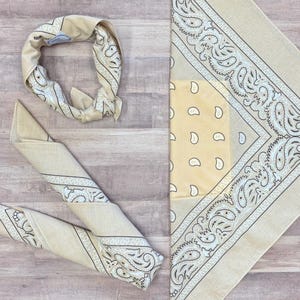 May include: A beige paisley bandana, folded and displayed in multiple ways. One is folded into a headband, another is rolled up, and the last is unfolded, showing the full paisley design. The bandana is made of 100% cotton.