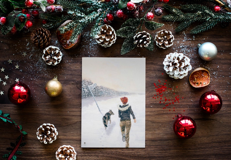 Op de afbeelding: Een kerstkaart met een besneeuwde wintersc&egrave;ne met een persoon en een hond die door een bos lopen. De kaart is omringd door dennenappels, hulst en ornamenten op een houten oppervlak.