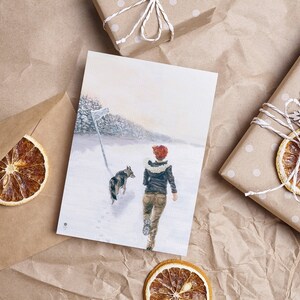 Op de afbeelding: Een aquarel schilderij van een persoon en een hond die door een besneeuwd bos lopen. De persoon draagt een rode hoed en een bruin jack. De hond is bruin en wit.