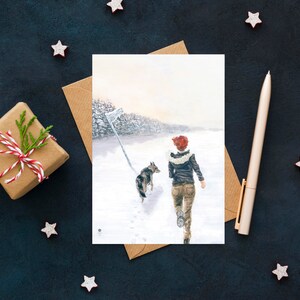 Op de afbeelding: Een aquarel schilderij van een vrouw die met een hond in een besneeuwd bos loopt. De vrouw draagt een rode hoed en een bruin jack. De hond is een grijs-witte husky.