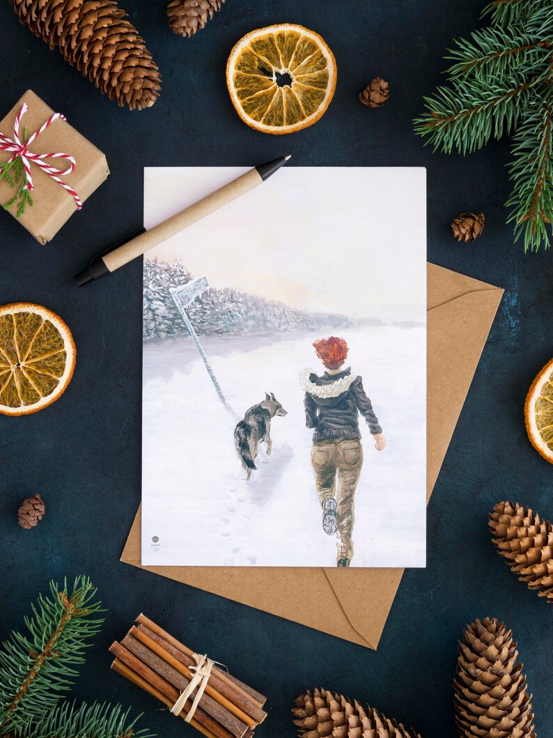 Op de afbeelding: Een aquarel illustratie van een persoon die een hond uitlaat in een besneeuwd bos. De persoon draagt een bruin jack en een rode sjaal. De hond is een bruin-witte hond. De illustratie is op een witte kaart met een bruine envelop.