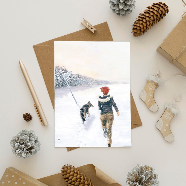 Op de afbeelding: Een vrouw met rood haar loopt door een besneeuwd landschap met haar hond. De vrouw draagt een bruin jack en een spijkerbroek. De hond is een grijs-witte wolfshond.