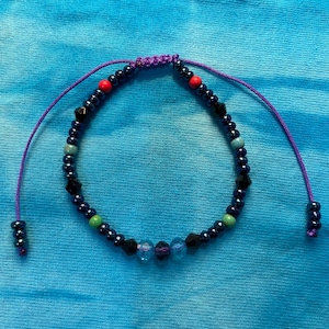 May include: A handmade bracelet with a sliding knot closure. The bracelet features dark blue beads, interspersed with red, green, and clear beads. The adjustable cord is purple, and the background is a blue tie-dye fabric.