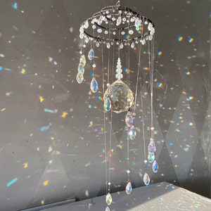 May include: A decorative crystal mobile with a circular top and multiple strands of clear crystals. The mobile features a large, faceted crystal ball and teardrop-shaped crystals, creating a shimmering effect. The crystals reflect light, producing a rainbow of colors.