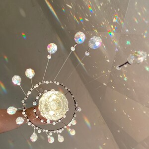 May include: A decorative crystal chandelier with a central globe and radiating crystal spheres. The chandelier features a double-ring design with smaller crystals and is held by a hand. The image is illuminated with rainbow light reflections.