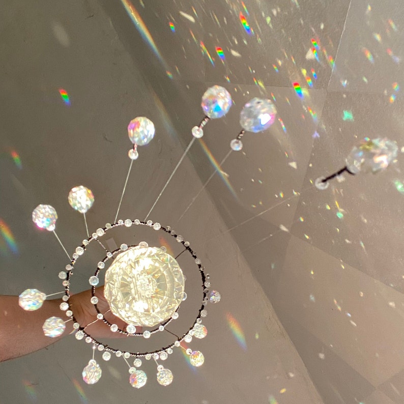 May include: A decorative crystal chandelier with a central globe and radiating crystal spheres. The chandelier features a double-ring design with smaller crystals and is held by a hand. The image is illuminated with rainbow light reflections.
