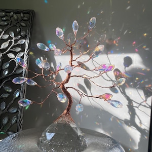 Op de afbeelding: Een decoratieve boomsculptuur met een koperkleurige draadstam en takken. De takken bevatten iriserende kristallen bladeren en staan op een heldere, facetgeslepen kristallen basis. De sculptuur wordt tentoongesteld op een reflecterend oppervlak.