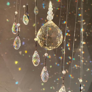 May include: A decorative crystal mobile with a large faceted sphere and teardrop-shaped crystals. The crystals are suspended from a dark ring and reflect light, creating a rainbow effect. The mobile is designed for home decor.