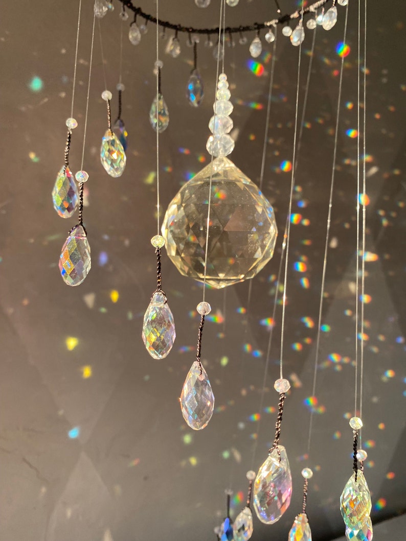 May include: A decorative crystal mobile with a large faceted sphere and teardrop-shaped crystals. The crystals are suspended from a dark ring and reflect light, creating a rainbow effect. The mobile is designed for home decor.