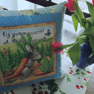 Könnte beinhalten: Ein quadratisches Zierkissen mit einem skurrilen Hasen- und Karotten-Design. Das Kissen hat einen hellblauen Rand mit weißen Punkten und ein zentrales Bild eines Hasen, der eine Karotte hält, mit dem Text "a big yummy carrot oh, the fun never ends."