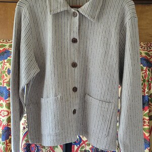 May include: A long-sleeved button-down shirt in a light beige fabric with thin vertical black stripes. The shirt features a classic collar, five dark brown buttons down the front, and two square patch pockets at the waist. An L.L.Bean label is visible.