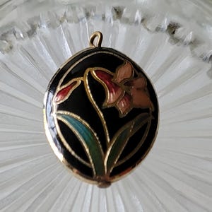 May include: An oval pendant featuring a black enamel background with a gold-toned metal outline. The design depicts a single flower with pink and red petals and green leaves, accented by a small gold-toned metal loop at the top for attachment.