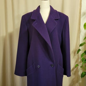 May include: A long, deep purple wool-blend coat featuring a structured collar, wide lapels, and a double-breasted front closure with two visible dark buttons. The coat has long sleeves and two slanted welt pockets positioned at the waist area.