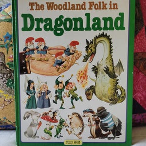 May include: A hardcover book titled The Woodland Folk in Dragonland by Tony Wolf. The cover features a large green dragon breathing fire, a giant hand holding small gnomes, and various woodland creatures including a rabbit, hedgehog, and bird.