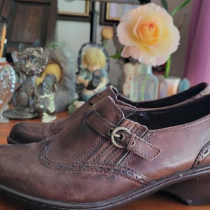 May include: A pair of dark brown leather slip-on shoes featuring a rounded toe, decorative brogue-style perforations along the seams, and a side strap with a small metallic buckle. The shoes have a low, stacked heel and a smooth, worn-in finish.