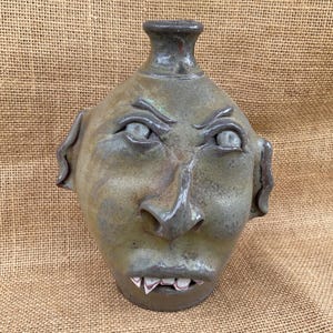 May include: A handcrafted ceramic jug with a face design. The jug features a detailed, expressive face with furrowed brows, a prominent nose, and a mouth revealing teeth. The color palette includes shades of green, gray, and brown.