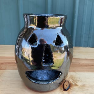 May include: A glossy black ceramic jack-o'-lantern candle holder with a wide, open mouth, a triangular nose, and two triangular eyes. The surface of the pottery is reflective, showing light highlights and a smooth, dark finish throughout.