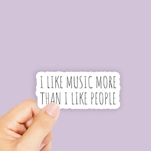 May include: White sticker with black text that reads "I like music more than I like people". A hand is holding the sticker.