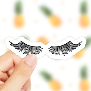 Könnte beinhalten: Ein weißer Aufkleber mit einer schwarzen Umrandung eines Paares Wimpern. Der Aufkleber wird von einer Hand gehalten.
