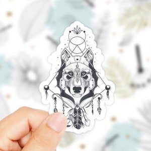 以下が含まれることがあります： 幾何学模様と羽根が付いたオオカミの頭を描いた白黒のステッカー。オオカミの頭は円と三角形に囲まれています。