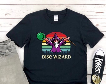 Puede incluir: Camiseta negra con un gráfico retro de un mago sosteniendo un disco de golf y una varita. El gráfico está sobre un fondo de puesta de sol con el texto "DISC WIZARD" debajo.