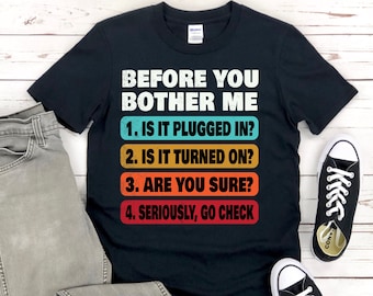 以下が含まれることがあります： 黒いTシャツに、白いグラフィックで「私を困らせる前に」と書いてあり、レトロなフォントで4つの質問がリストされています。「1. 接続されていますか？ 2. 電源が入っていますか？ 3. 確実ですか？ 4. 本気で確認してください。」