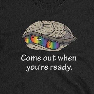 May include: Black t-shirt with a cartoon illustration of a turtle shell with rainbow colored turtles inside. The text "Come out when you're ready." is printed below the image.