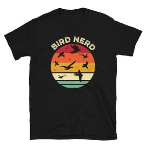 May include: Black t-shirt with a vintage-style graphic of a circle with a red, orange, yellow, and green background. The circle has silhouettes of birds flying in it. The text "Bird Nerd" is above the circle.