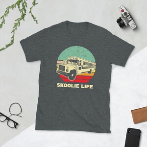 May include: A dark heather grey t-shirt with a vintage-style illustration of a yellow school bus driving through a sunset. The text "SKOOLIE LIFE" is printed below the bus.