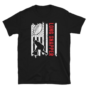 May include: Black t-shirt featuring a distressed American flag design with a football and a football player silhouette. The text "LONG SNAPPER" is printed vertically in red. The shirt is made of a soft, comfortable material.