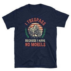 Pu&ograve; includere: T-shirt blu navy con una grafica di funghi spugnole e la scritta "I TRESSPASS BECAUSE I HAVE NO MORELS". Il design include un cerchio a strisce rosse e bianche e dettagli verdi.