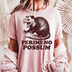 May include: A light pink t-shirt featuring a detailed illustration of an opossum with its mouth open, and the text "PERIMENO POSSUM" in a bold, dark brown font. The opossum graphic is centered on the shirt.