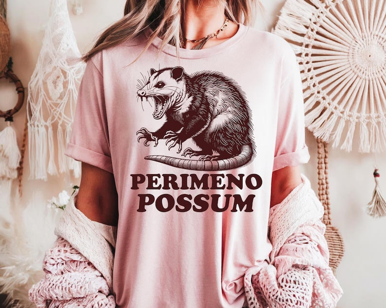 May include: A light pink t-shirt featuring a detailed illustration of an opossum with its mouth open, and the text "PERIMENO POSSUM" in a bold, dark brown font. The opossum graphic is centered on the shirt.