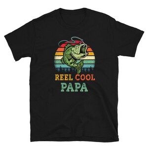 Pu&ograve; includere: Maglietta nera con un'illustrazione in stile retr&ograve; di un pesce su un amo da pesca. Il testo "Reel Cool Papa" &egrave; stampato sotto l'immagine.