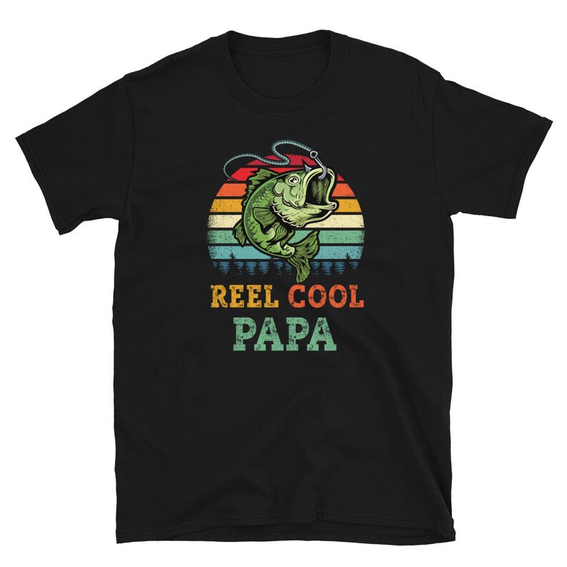Pu&ograve; includere: Maglietta nera con un'illustrazione in stile retr&ograve; di un pesce su un amo da pesca. Il testo "Reel Cool Papa" &egrave; stampato sotto l'immagine.
