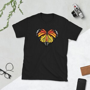Puede incluir: Camiseta negra con un dise&ntilde;o de mariposa en forma de coraz&oacute;n. La mariposa tiene alas naranjas, amarillas y negras.