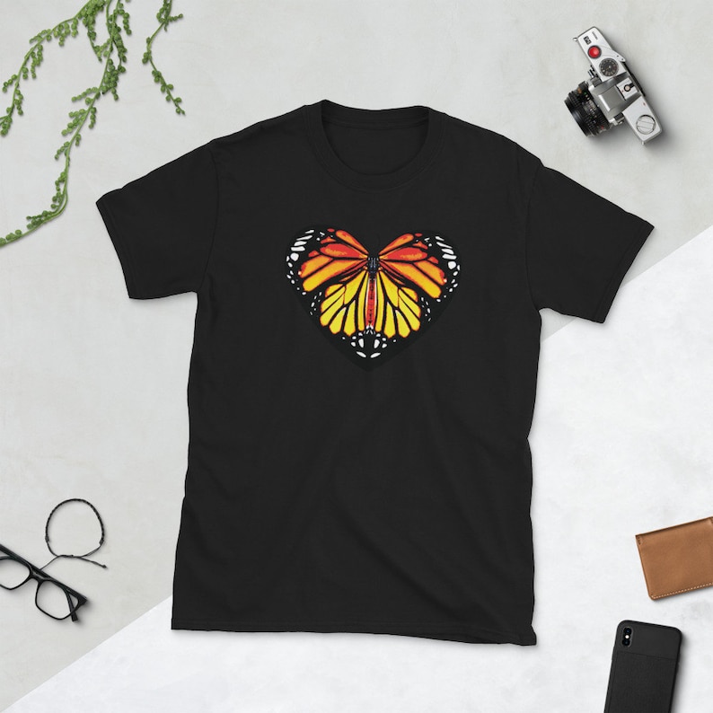 Puede incluir: Camiseta negra con un dise&ntilde;o de mariposa en forma de coraz&oacute;n. La mariposa tiene alas naranjas, amarillas y negras.
