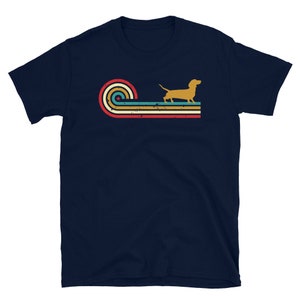 K&ouml;nnte beinhalten: Ein marineblaues T-Shirt mit einem Retro-Design, das eine Dackel-Silhouette und bunte Streifen zeigt. Das Design erinnert an die &Auml;sthetik der 1970er Jahre.